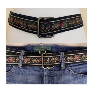 Può includere: Una cintura intrecciata nera e dorata con un motivo floreale. La cintura ha una fibbia argentata con il testo "LRL LAUREN JEANS CO. RALPH LAUREN" su di essa.