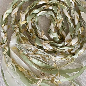 Peut inclure: Une coiffe tressée composée de rubans ivoire, vert clair et dorés. Les rubans sont entrelacés avec de la dentelle délicate et ornés d'accents de feuilles d'or, créant un design élégant inspiré de la nature. Idéal pour les mariages.