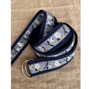 May include: A gray and navy blue fabric belt with a floral pattern. The belt has a gold metal buckle.