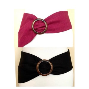 May include: Two fabric belts with a large, round, wooden buckle. The top belt is a deep pink color and the bottom belt is black.