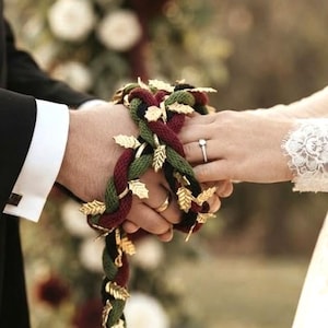 May include: A handfasting cord featuring braided dark red, forest green, and black rope strands. The cord is adorned with numerous small, metallic gold-colored leaf charms attached throughout the length of the braided rope design.