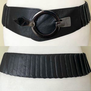 May include: Black wide belt with a large, circular silver-tone buckle and a smaller rectangular clasp. The belt has a textured appearance and a pleated design on the sides. The belt is shown in two views.