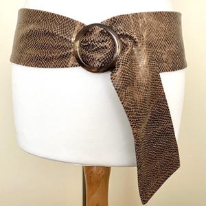 Puede incluir: Cinturón ancho marrón con estampado de piel de serpiente y una hebilla grande y redonda de carey. El cinturón está envuelto alrededor de una forma blanca, con un extremo colgando. Accesorio de moda.