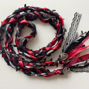 May include: A decorative hair accessory with a braided design. It features red satin ribbon, black lace, and silver accents. The accessory is adorned with small, dark leaves and decorative silver buttons. The overall aesthetic is gothic and romantic.