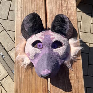 Op de afbeelding: Een handgemaakt dierenmasker met een paars en wit gevlekt gezicht, zwarte oren en paarse ogen. Het masker heeft een harige textuur en ligt op een houten oppervlak. Het masker is ontworpen om op een hyena te lijken.
