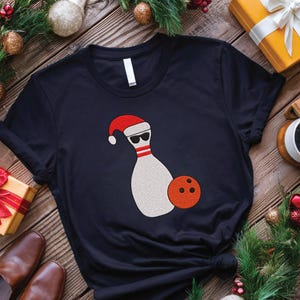 May include: Black t-shirt featuring a bowling pin wearing a Santa hat and sunglasses, with an orange bowling ball. The shirt is surrounded by Christmas decorations and gifts, perfect for the holidays.