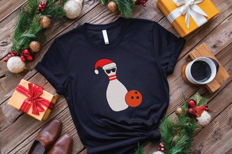 May include: Black t-shirt featuring a bowling pin wearing a Santa hat and sunglasses, with an orange bowling ball. The shirt is surrounded by Christmas decorations and gifts, perfect for the holidays.