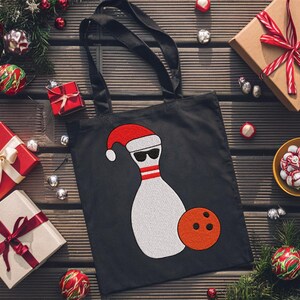 May include: Black tote bag featuring a bowling pin wearing a Santa hat and sunglasses, with an orange bowling ball. The bag is surrounded by Christmas decorations and wrapped gifts.