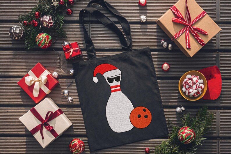 May include: Black tote bag featuring a bowling pin wearing a Santa hat and sunglasses, with an orange bowling ball. The bag is surrounded by Christmas decorations and wrapped gifts.