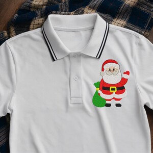 Puede incluir: Polo blanco con ribetes negros en el cuello y las mangas. La camisa presenta un dise&ntilde;o bordado de Pap&aacute; Noel en la parte delantera, sosteniendo un saco verde. El dise&ntilde;o de Pap&aacute; Noel incluye colores rojo, blanco, negro y dorado.