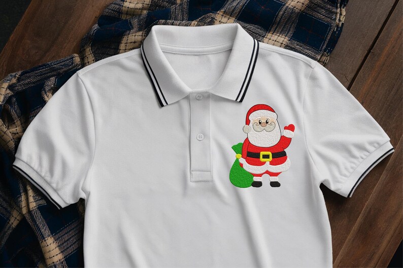 Puede incluir: Polo blanco con ribetes negros en el cuello y las mangas. La camisa presenta un dise&ntilde;o bordado de Pap&aacute; Noel en la parte delantera, sosteniendo un saco verde. El dise&ntilde;o de Pap&aacute; Noel incluye colores rojo, blanco, negro y dorado.
