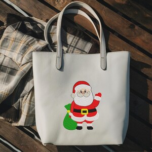 Puede incluir: Un bolso tote de color gris claro con un dise&ntilde;o de Pap&aacute; Noel. Pap&aacute; Noel lleva un traje rojo, un cintur&oacute;n negro con hebilla dorada y un gorro rojo. Sostiene una bolsa verde. Una bufanda a cuadros est&aacute; al lado de la bolsa.