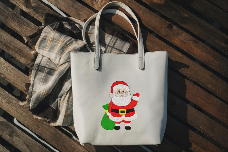 Puede incluir: Un bolso tote de color gris claro con un dise&ntilde;o de Pap&aacute; Noel. Pap&aacute; Noel lleva un traje rojo, un cintur&oacute;n negro con hebilla dorada y un gorro rojo. Sostiene una bolsa verde. Una bufanda a cuadros est&aacute; al lado de la bolsa.