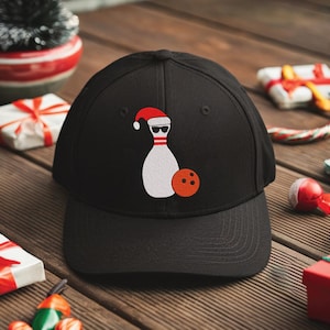 May include: Black baseball cap featuring a white bowling pin wearing a Santa hat and sunglasses, with an orange bowling ball. The cap is surrounded by Christmas-themed decorations on a wooden surface.