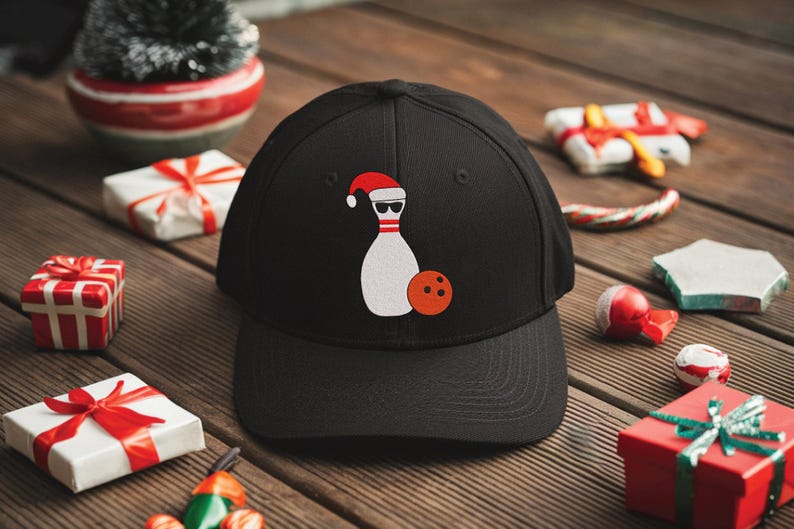 May include: Black baseball cap featuring a white bowling pin wearing a Santa hat and sunglasses, with an orange bowling ball. The cap is surrounded by Christmas-themed decorations on a wooden surface.