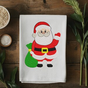 Puede incluir: Toalla blanca con tem&aacute;tica navide&ntilde;a con un dise&ntilde;o bordado de Pap&aacute; Noel. Pap&aacute; Noel est&aacute; representado con su traje rojo tradicional con cintur&oacute;n negro y un saco verde de regalos. Sobre una superficie de madera.