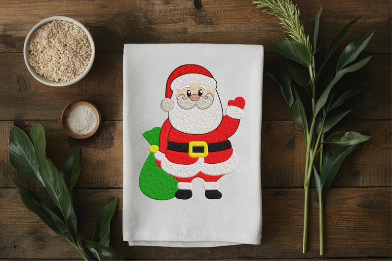 Puede incluir: Toalla blanca con tem&aacute;tica navide&ntilde;a con un dise&ntilde;o bordado de Pap&aacute; Noel. Pap&aacute; Noel est&aacute; representado con su traje rojo tradicional con cintur&oacute;n negro y un saco verde de regalos. Sobre una superficie de madera.