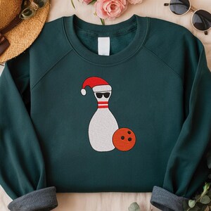 May include: Dark teal sweatshirt with a graphic of a bowling pin wearing sunglasses and a Santa hat, with an orange bowling ball. The sweatshirt has a crew neck and long sleeves with gray cuffs. A straw hat and sunglasses are in the background.