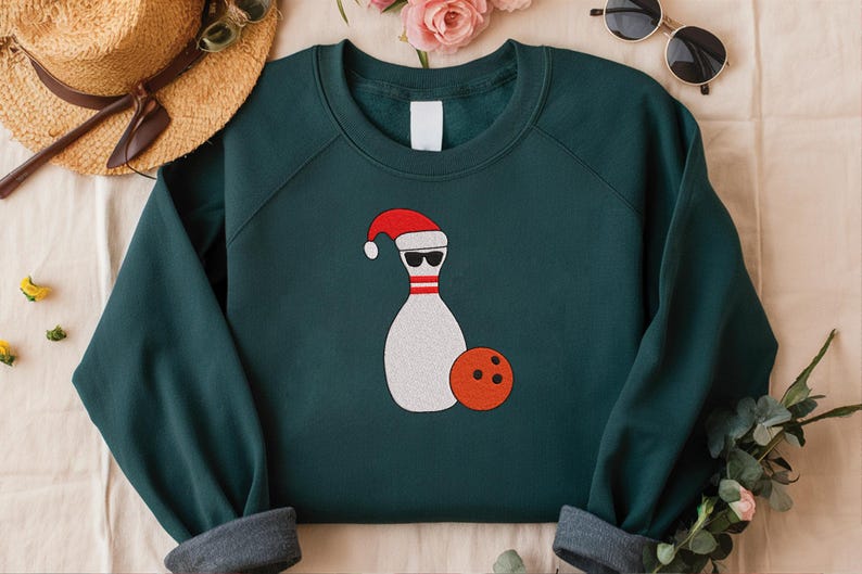 May include: Dark teal sweatshirt with a graphic of a bowling pin wearing sunglasses and a Santa hat, with an orange bowling ball. The sweatshirt has a crew neck and long sleeves with gray cuffs. A straw hat and sunglasses are in the background.