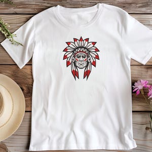 Pode incluir: Uma t-shirt branca de manga curta com um gráfico central de uma pessoa a usar um cocar de penas tradicional. A ilustração utiliza cores preto, branco e vermelho para representar o rosto e as penas detalhadas do adorno de cabeça.