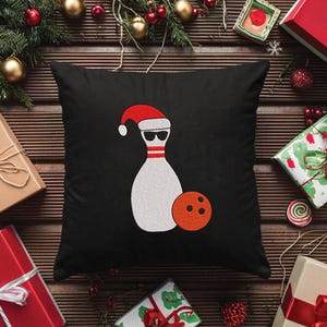 May include: Black square pillow featuring a bowling pin wearing a Santa hat and sunglasses, with an orange bowling ball. The pillow is surrounded by wrapped gifts, Christmas ornaments, and greenery on a wooden surface.