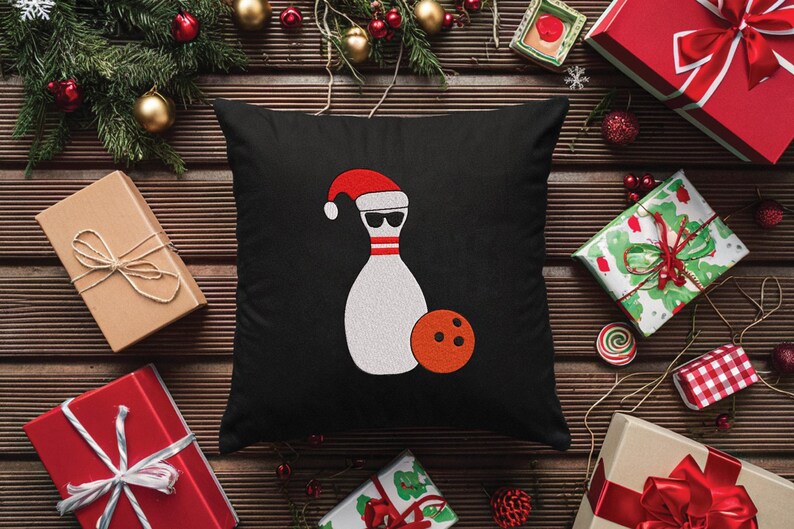 May include: Black square pillow featuring a bowling pin wearing a Santa hat and sunglasses, with an orange bowling ball. The pillow is surrounded by wrapped gifts, Christmas ornaments, and greenery on a wooden surface.
