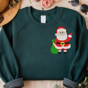 Puede incluir: Una sudadera verde oscuro con un dise&ntilde;o de Pap&aacute; Noel. Pap&aacute; Noel lleva un traje rojo, un cintur&oacute;n negro con hebilla dorada y barba blanca. Sostiene un saco verde. La sudadera tiene cuello redondo y mangas largas.