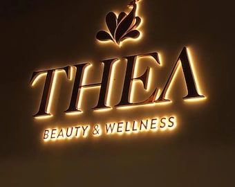 Könnte beinhalten: Ein goldfarbenes Schild mit dem Wort "THEA" in großen Buchstaben und den Worten "BEAUTY & WELLNESS" in kleineren Buchstaben darunter. Das Schild ist mit warmweißen Lichtern beleuchtet.