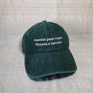 May include: A dark green baseball cap featuring white embroidered text on the front that reads "nascida para viajar, forçada a estudar". The cap has a structured crown, a curved brim, and a classic six-panel design with a button on the top.
