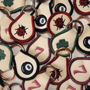 Peut inclure: Un assortiment de porte-clés brodés avec divers motifs. Certains porte-clés présentent une coccinelle, le chiffre 7, une boule numéro 8 et un trèfle à quatre feuilles. Chaque porte-clés a un anneau métallique pour l'attache.