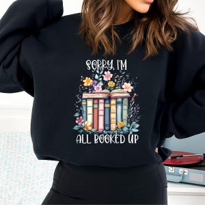 May include: Black sweatshirt with a graphic of a stack of books with flowers and the text "Sorry, I'm all booked up".