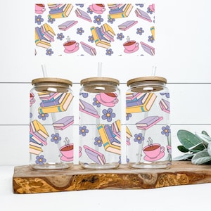 May include: Three clear glass tumblers with wooden lids and straws. Each tumbler features a colorful pattern of stacked books, coffee cups, and flowers. The tumblers are sitting on a wooden surface.