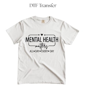 May include: White t-shirt with the text "MENTAL HEALTH matters" in a black, stylized font. The phrase "ALL DAY EVERY DAY" is below the main text. The words are enclosed in a black rectangle with hearts.