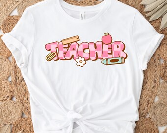 May include: White t-shirt with the word "TEACHER" in pink and white checked letters. The letters are surrounded by small school-themed icons, including a ruler, a paperclip, a flower, an apple, and a crayon.