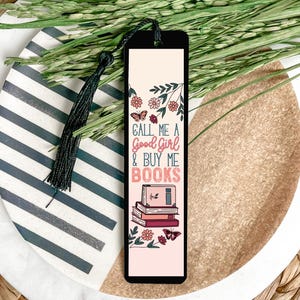 May include: A pink and black bookmark with a floral design and the text "Call me a good girl & buy me books". The bookmark features a stack of books with a butterfly on top.