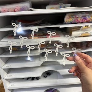 Puede incluir: Adornos blancos en forma de lazo con cuentas colgantes de perlas se muestran en una lámina transparente. Una mano con manicura morada sostiene algunos de los lazos. El fondo muestra un mueble de almacenamiento blanco con suministros de artesanía.