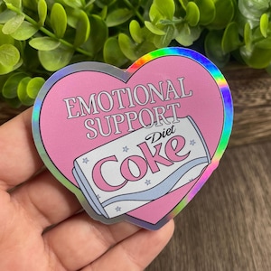 Może przedstawiać: Różowa naklejka w kształcie serca z holograficznym wykończeniem. Naklejka zawiera tekst "Emotional Support Diet Coke" ze stylizowaną puszką Diet Coke.