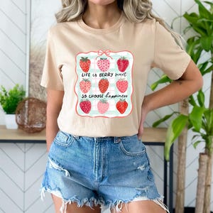 以下が含まれることがあります： イチゴのグラフィックがプリントされたベージュのTシャツ。グラフィックには9つのイチゴと「Life is berry sweet so choose happiness」の文字が描かれています。デニムショーツと合わせています。