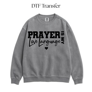 以下が含まれることがあります： 「PRAYER is MY Love language」の黒い文字がプリントされたグレーのクルーネックのスウェットシャツ。 「Love language」という言葉の下には小さな黒いハートがあります。 画像の上部には「DTF Transfer」という文字があります。