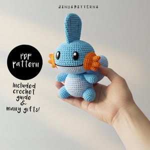 Pode incluir: Um personagem Pokemon de crochê azul e laranja, Froakie, é segurado na mão de uma pessoa. A imagem inclui o texto "PDF pattern" e "Included crochet guide & many gifts!"