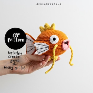 Puede incluir: Un pez de ganchillo naranja y amarillo con aletas blancas y una boca rosa. El pez está siendo sostenido por una mano. El texto "PDF pattern" está en un círculo negro y el texto "Included crochet guide & many gifts!" está debajo del círculo.