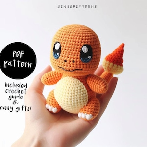 May include: A crocheted orange and yellow Pokemon character with a tail. The character has large blue eyes and a small mouth. The character is being held in a person's hand.