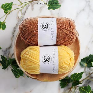 Op de afbeelding: Twee bollen garen, een bruin en een geel, met etiketten waarop "Sweet Candy" staat.
