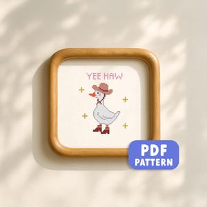 May include: A framed cross-stitch pattern featuring a white goose wearing a cowboy hat and boots. The text "YEE HAW" is above the goose. The frame is made of wood, and a blue PDF pattern button is in the lower right corner.