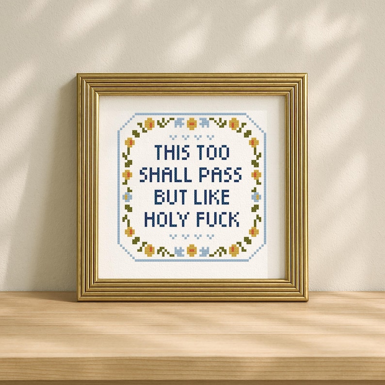May include: A framed cross-stitch artwork with the text "THIS TOO SHALL PASS BUT LIKE HOLY FUCK" in blue. The design is surrounded by a floral border in yellow and blue, set against a cream background. The frame is gold.