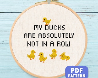 Puede incluir: Un bastidor de bordado de punto de cruz con el texto "MY DUCKS ARE ABSOLUTELY NOT IN A ROW" en negro. El diseño incluye patitos amarillos pixelados. Un botón de patrón PDF azul está en la esquina inferior derecha.
