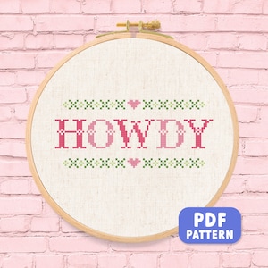 May include: A cross-stitch embroidery hoop with the word "HOWDY" in pink, surrounded by green and pink cross-stitch patterns. The design is on a cream-colored fabric, set in a wooden hoop, and against a pink brick wall. A PDF pattern button is in the bottom right corner.