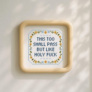 May include: Square framed cross-stitch art with the text "THIS TOO SHALL PASS BUT LIKE HOLY FUCK". The artwork is surrounded by a floral border in yellow and blue, set against a white background.