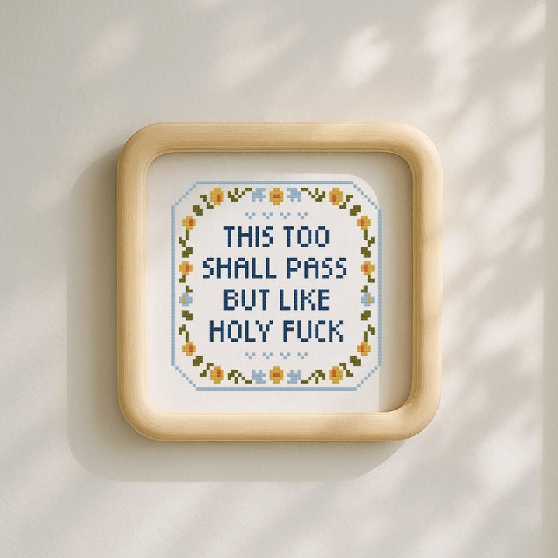 May include: Square framed cross-stitch art with the text "THIS TOO SHALL PASS BUT LIKE HOLY FUCK". The artwork is surrounded by a floral border in yellow and blue, set against a white background.