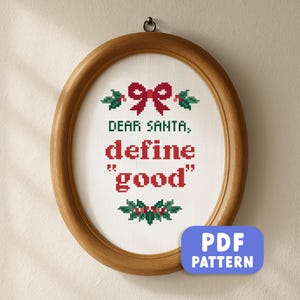May include: Oval wooden framed cross-stitch with the text "DEAR SANTA, define "good"" in red. The design includes a red bow and holly accents. A blue PDF pattern button is in the lower right corner.